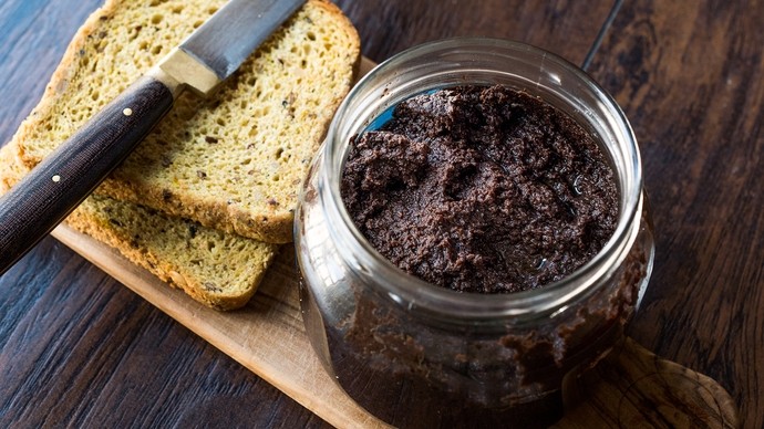 An open jar of olive tapenade next to a knife on two pieces of toasted bread