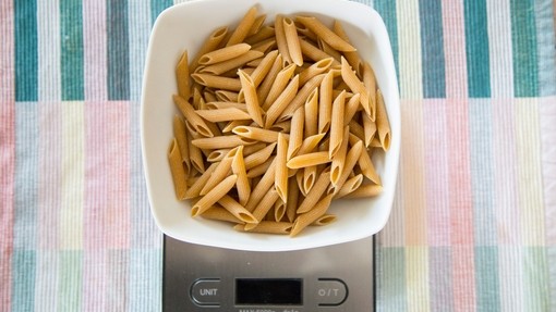 A white bowl full of uncooked pasta on a silver digital food scale, laid on a striped tablecloth.