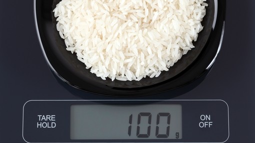 A black bowl of dried rice being measured on a digital food scale. The screen says 100g.