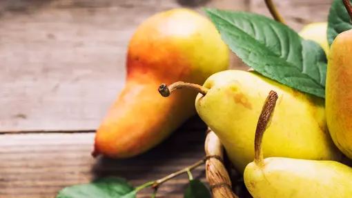 A photo of green and peach coloured pears spilling onto a dark wooden table top with green leaves scattered decoratively