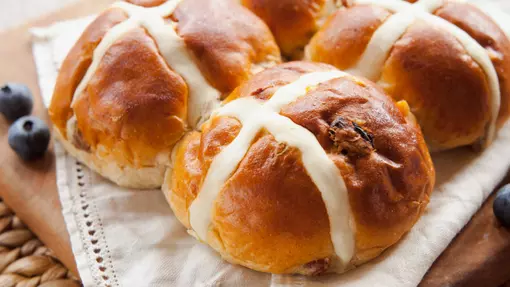 A wooden tray full of hot cross buns, blueberries are scattered around the edge