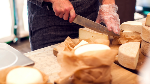 A wooden table with different cheese scattered on it. A white hand holding a knife is cutting a wedge of cheese on the table
