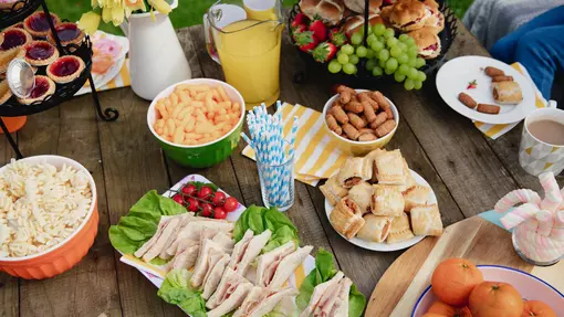 An outdoor table with picnic food such as pasta, crisps, sausage rolls and sandwiches