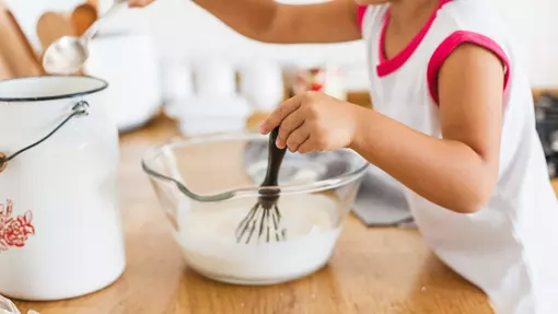 a baking bowl with white mix, a toddler is holding a whisk in the bowl and has a spoon in the other hand dipping in a pot