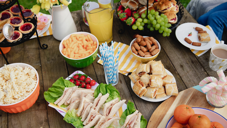 An outdoor table with picnic food such as pasta, crisps, sausage rolls and sandwiches