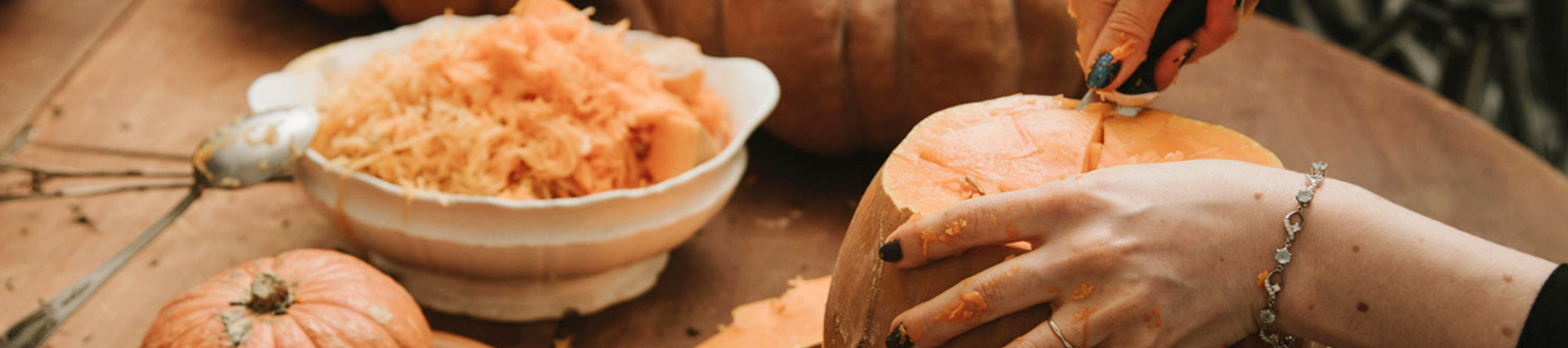 A photo of two people's hands cutting and gutting a couple of different sized orange pumpkins on a brown surface. A bowl of pumpkin flesh is also on the table.